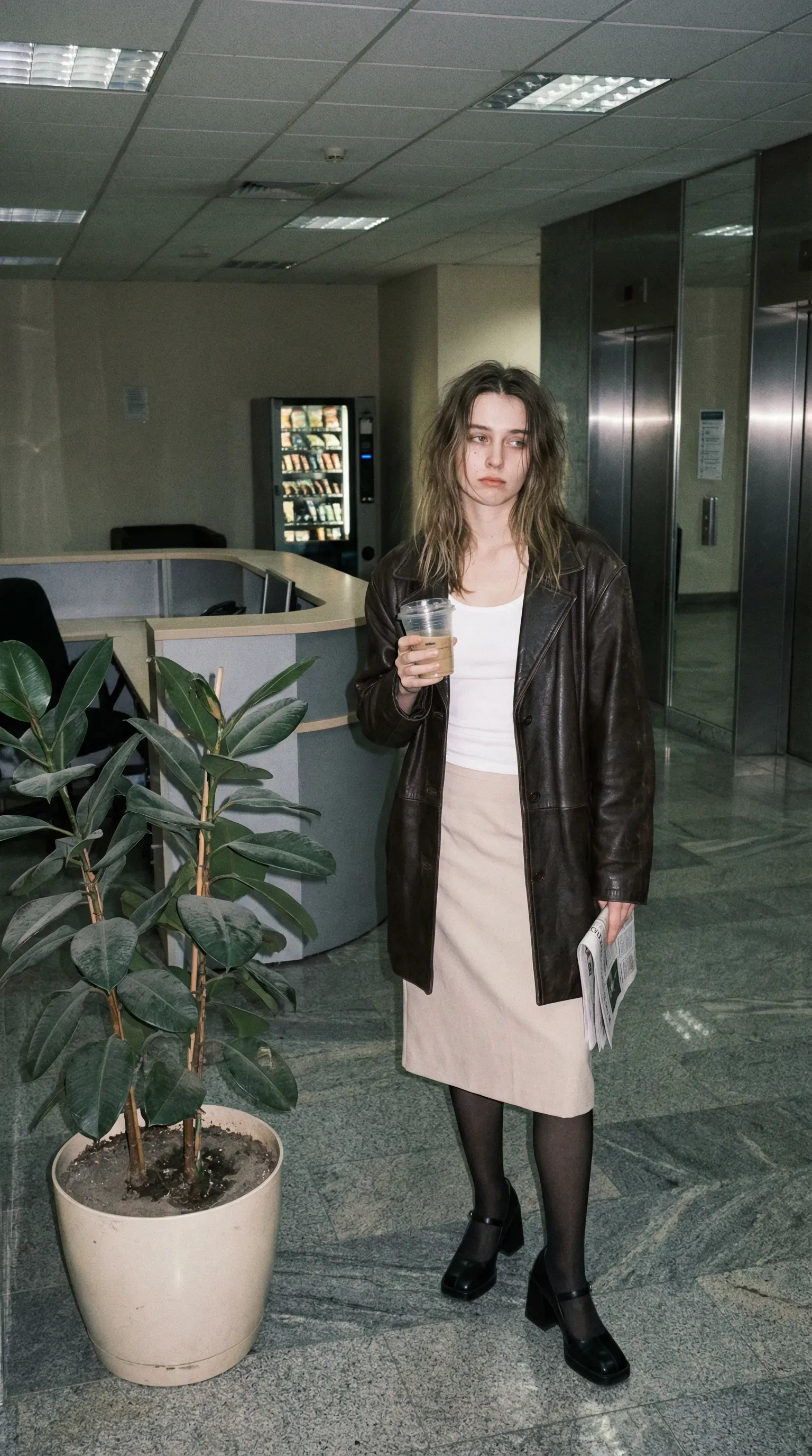 Casual smartphone camera photo, full-body portrait of a young woman alone in an office building lobby after hours. Flat interior lighting with slight flash, cool beige and grey palette, low contrast, mild digital grain.
Subject: Standing beside a huge potted plant, wearing a dark brown leather coat over a white tank and pale knee-length skirt, with black tights and square-toe heels. She holds a plastic takeaway coffee cup and a folded newspaper under one arm. Hair slightly undone, expression tired and unreadable.
Background: Empty reception desk, glossy stone floor, mirrored elevator doors, and a vending machine glowing in one corner. The plant is oversized and slightly dusty, giving the scene a strange corporate melancholy. Quiet fashion image, lonely city-girl realism, early-2000s candid energy.