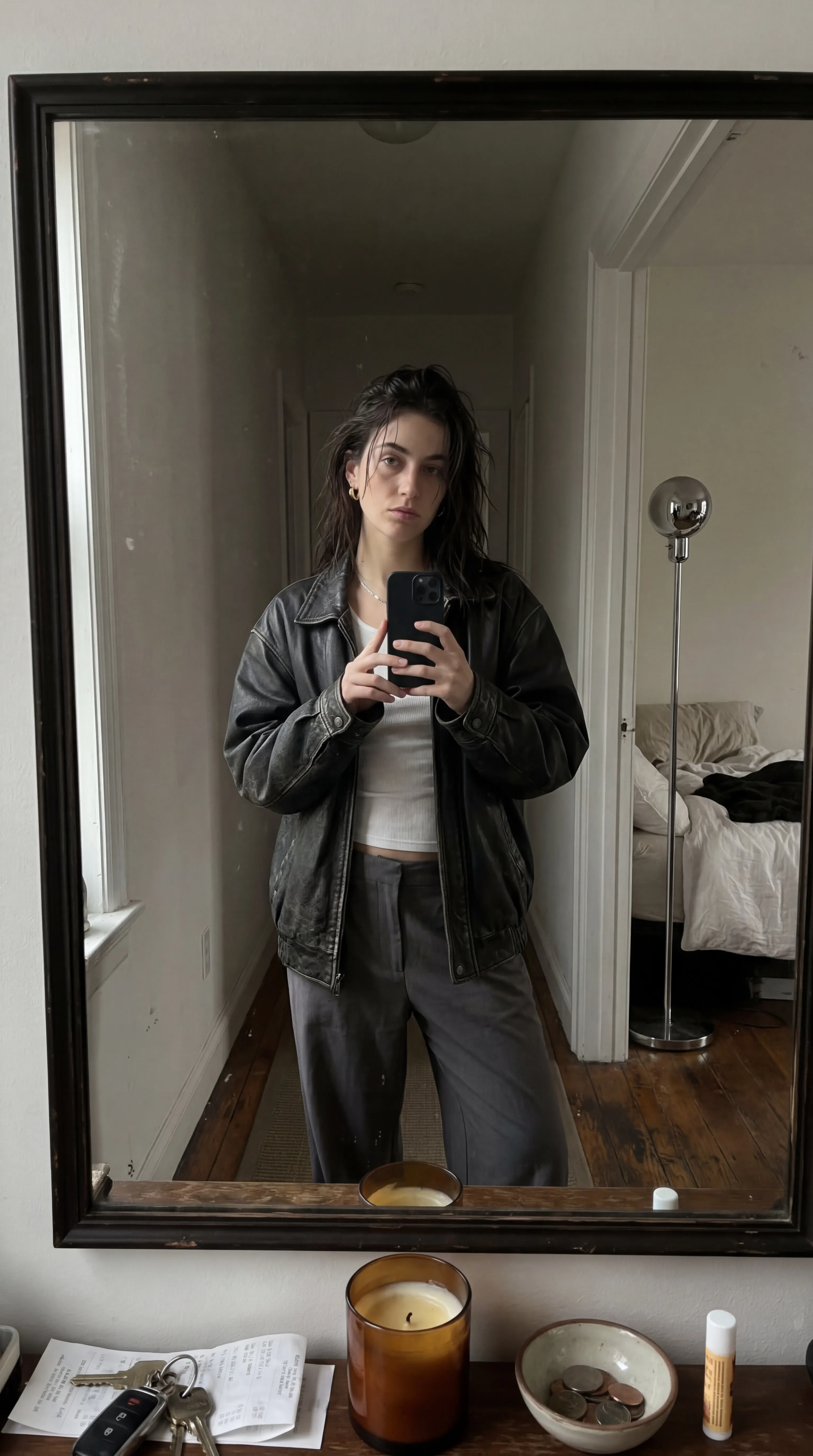 Casual unedited smartphone mirror selfie, dim late-afternoon apartment light, muted neutral palette with soft beige, washed black, and silvery grey tones. Slight digital grain, low contrast, candid but composed aesthetic.
Subject: A young woman standing in a narrow hallway mirror, holding a dark phone loosely at chest height. She wears an oversized black leather jacket over a thin white ribbed tank and loose charcoal trousers sitting low on the hips. Hair messy, slightly damp, with a tired expensive look.
Foreground: The edge of a cluttered console table with keys, receipts, a half-burned candle, and a shallow metal bowl.
Background: A partially open door reveals a bed with crumpled white sheets and a chrome floor lamp. White walls, dark wood floor, no dramatic styling, intimate off-duty fashion mood.