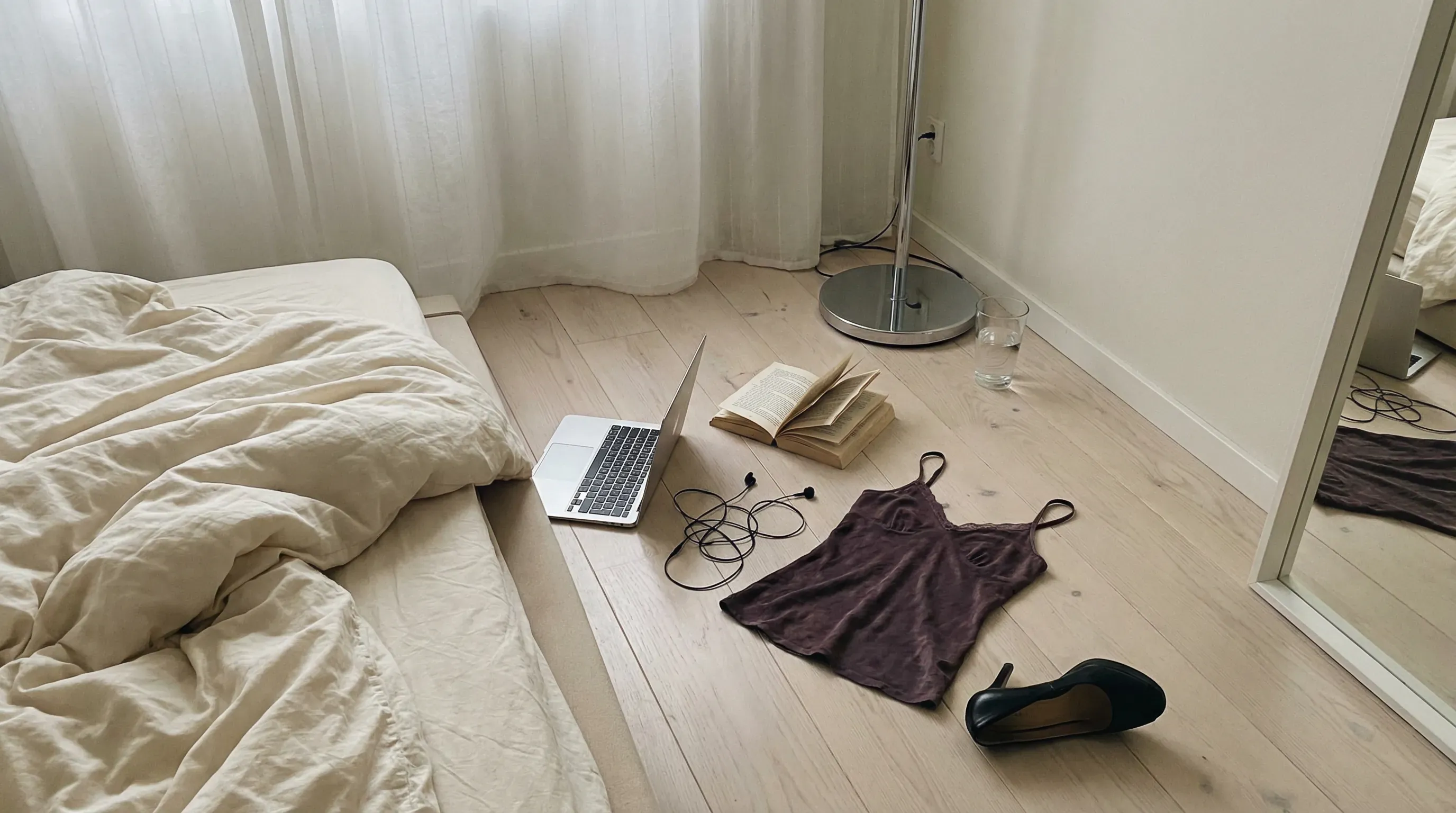 Casual unedited iPhone snapshot, morning bedroom floor scene, flat daylight, soft desaturated palette of cream, silver, faded plum, and black. No people visible, only traces of someone just there.
Scene: A rumpled duvet spilling off a low bed onto pale wood floors. Nearby: a half-open silver laptop, a paperback with folded pages, wired earphones, a dark red camisole, and one black heel turned upside down.
Side area: A cylindrical chrome lamp base and a glass of water placed directly on the floor instead of a bedside table.
Background: Sheer curtains, plain wall, edge of a mirror reflecting only light and part of the bed. Quiet intimate mess, tasteful and unforced, emotionally suggestive without being staged.