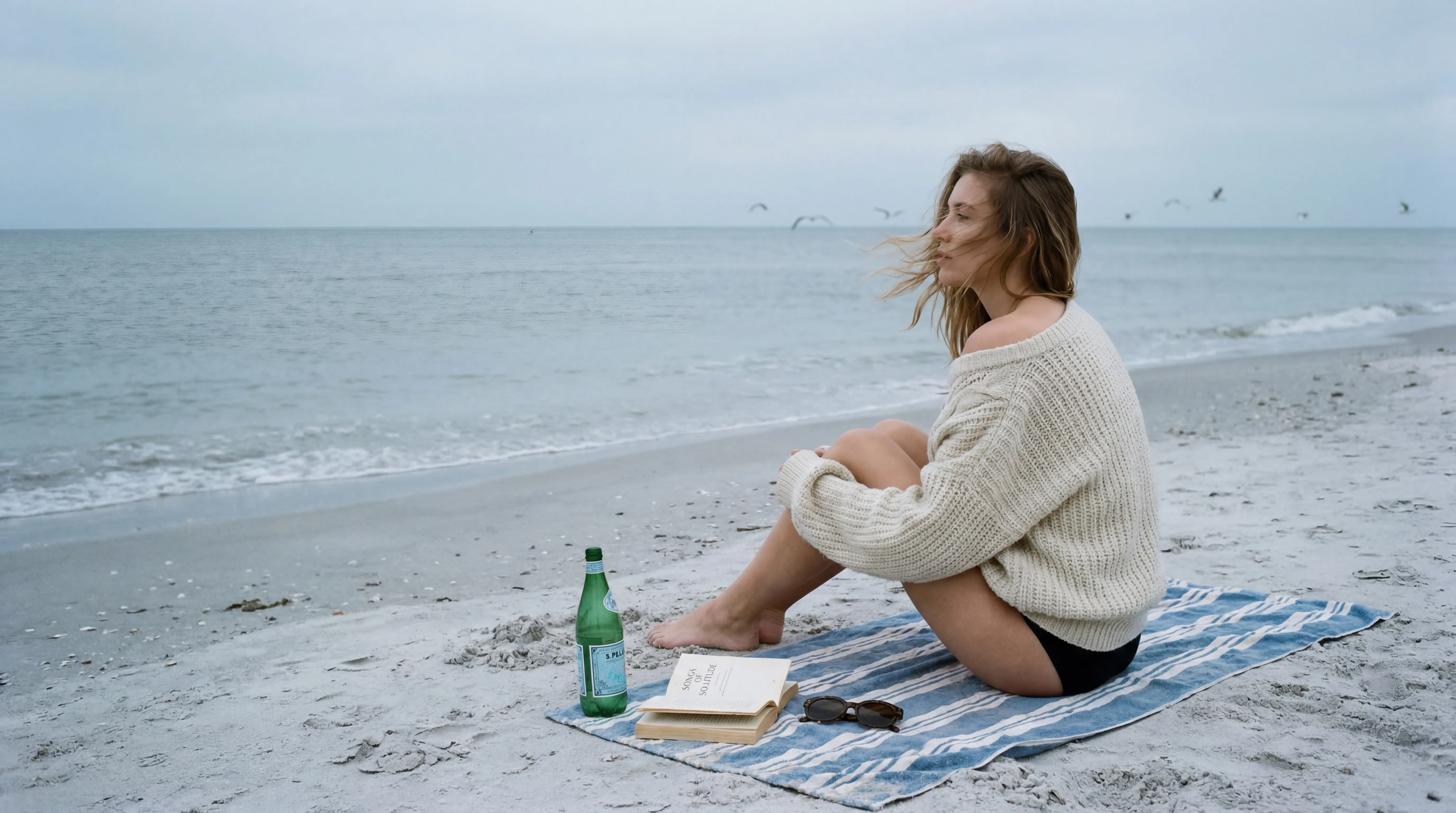 Editorial 35mm film photography, overcast beach light, extremely soft shadows, pale cool sand tones, faded ocean blues, and heavy film grain. Quiet melancholy with understated luxury.
Subject: A woman sitting alone on a striped towel near the shoreline, knees bent, wearing an oversized cream fisherman sweater slipping off one shoulder and black bikini bottoms. Wind pushing strands of hair across her face.
Objects: Beside her sits an open paperback, dark sunglasses, and a half-empty green glass bottle of mineral water.
Background: Flat grey-blue sea, empty horizon, a few distant seagulls, no other people visible. Minimal coastal solitude, cinematic stillness, contemplative summer sadness.