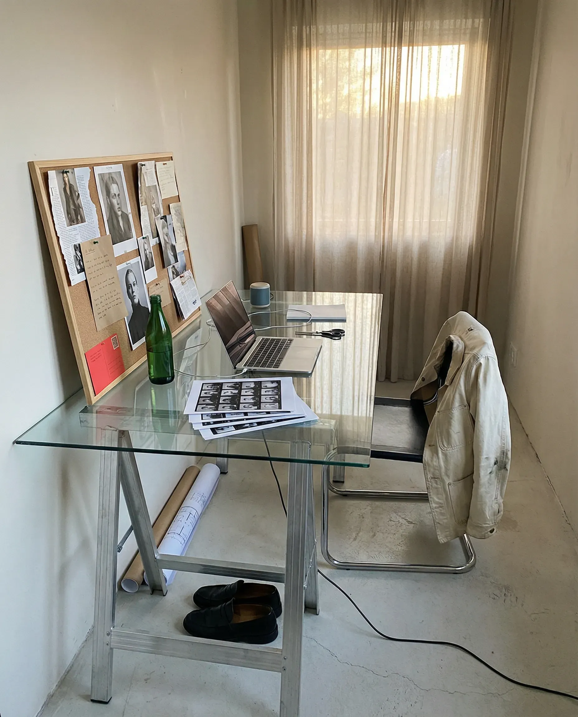 Casual unedited smartphone snapshot, late afternoon natural light, slightly desaturated neutral palette with muted tobacco brown, chalk white, faded steel blue, and brushed aluminum. A narrow art-school style studio space with a calm, expensive, semi-temporary feel.
Foreground: A large sheet of glass laid across two raw aluminum trestles as a makeshift desk. On top: an open silver laptop, a stack of loose printed contact sheets, silver scissors, a green glass bottle, and a tiny wired desk speaker.
Left: A freestanding cork board leaning against the wall, pinned with torn magazine pages, monochrome portraits, handwritten notes, and one bright red postcard.
Right: A tubular chrome dining chair with a cream canvas jacket slung over the back. Under the desk sits a pair of black square-toe loafers and a rolled blueprint.
Background: Plain off-white wall, pale concrete floor, one trailing cable visible, sheer curtains glowing softly from an unseen window. Amateur iPhone realism, understated creative-director workspace mood.