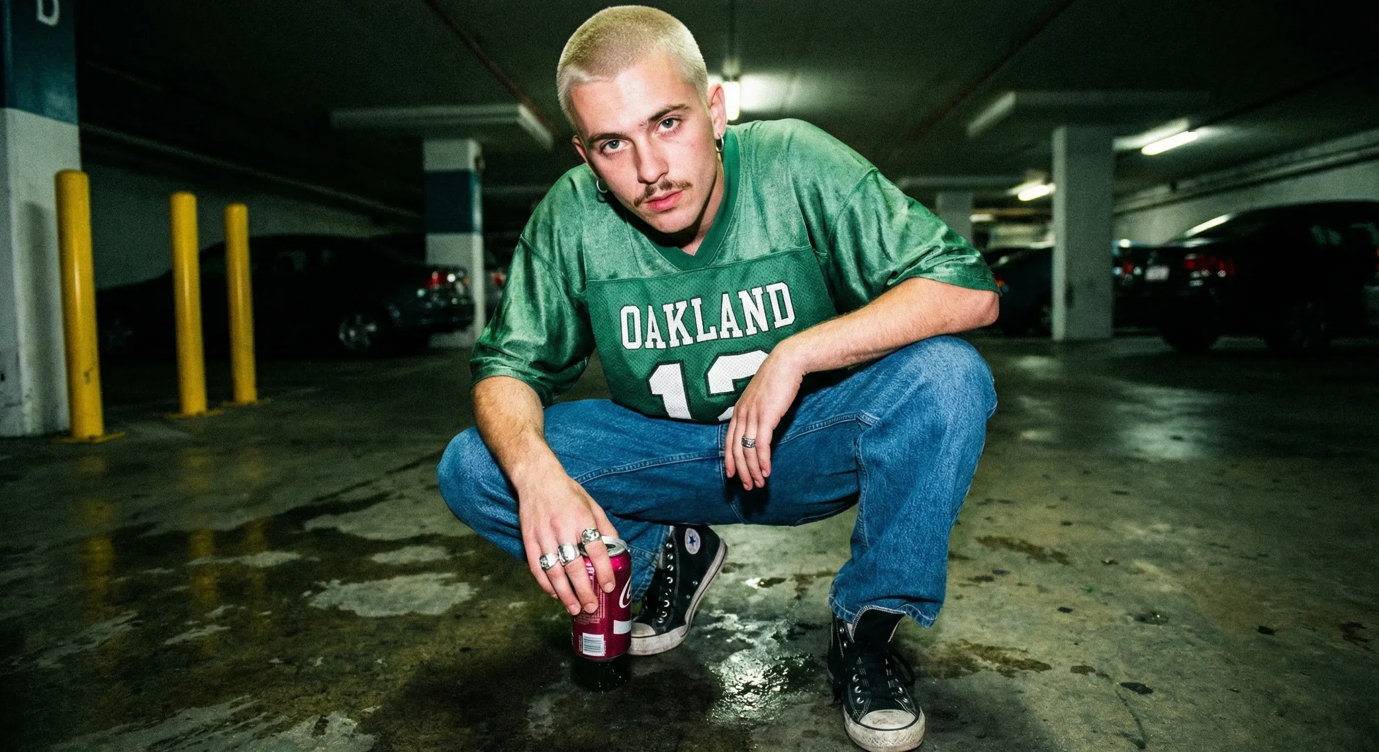 90s fashion snapshot, harsh direct flash, wide-angle lens distortion, saturated but slightly dirty urban color palette. Energetic, imperfect, street-cast mood.
Subject: A young man crouching in a parking garage, elbows on knees, staring directly into camera. Bleached buzzcut, thin mustache, silver hoop earring. Wearing a faded green football jersey, oversized jeans, and scuffed black sneakers.
Details: One hand holds a cherry-red soda can, the other hangs loosely with rings catching flash. Oily concrete floor reflects small patches of light.
Background: Yellow safety bollards, painted parking lines, shadowy cars barely visible. Raw youth-culture editorial feel.