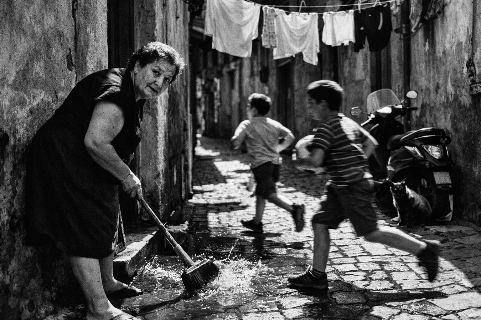 35mm documentary-style street photograph, black and white, strong noon light, crisp contrast, dynamic candid movement.
Scene: A narrow southern European alley with laundry strung overhead and sharp slices of sunlight on cobblestones. In the foreground, an older woman in a dark dress sweeps water from her doorstep while glancing toward the camera.
Midground: Two boys run past carrying a football, slightly blurred in motion. A cat sits in shadow beneath a scooter.
Mood: Everyday poetry, lived-in urban texture, timeless human warmth, honest street-photography composition.