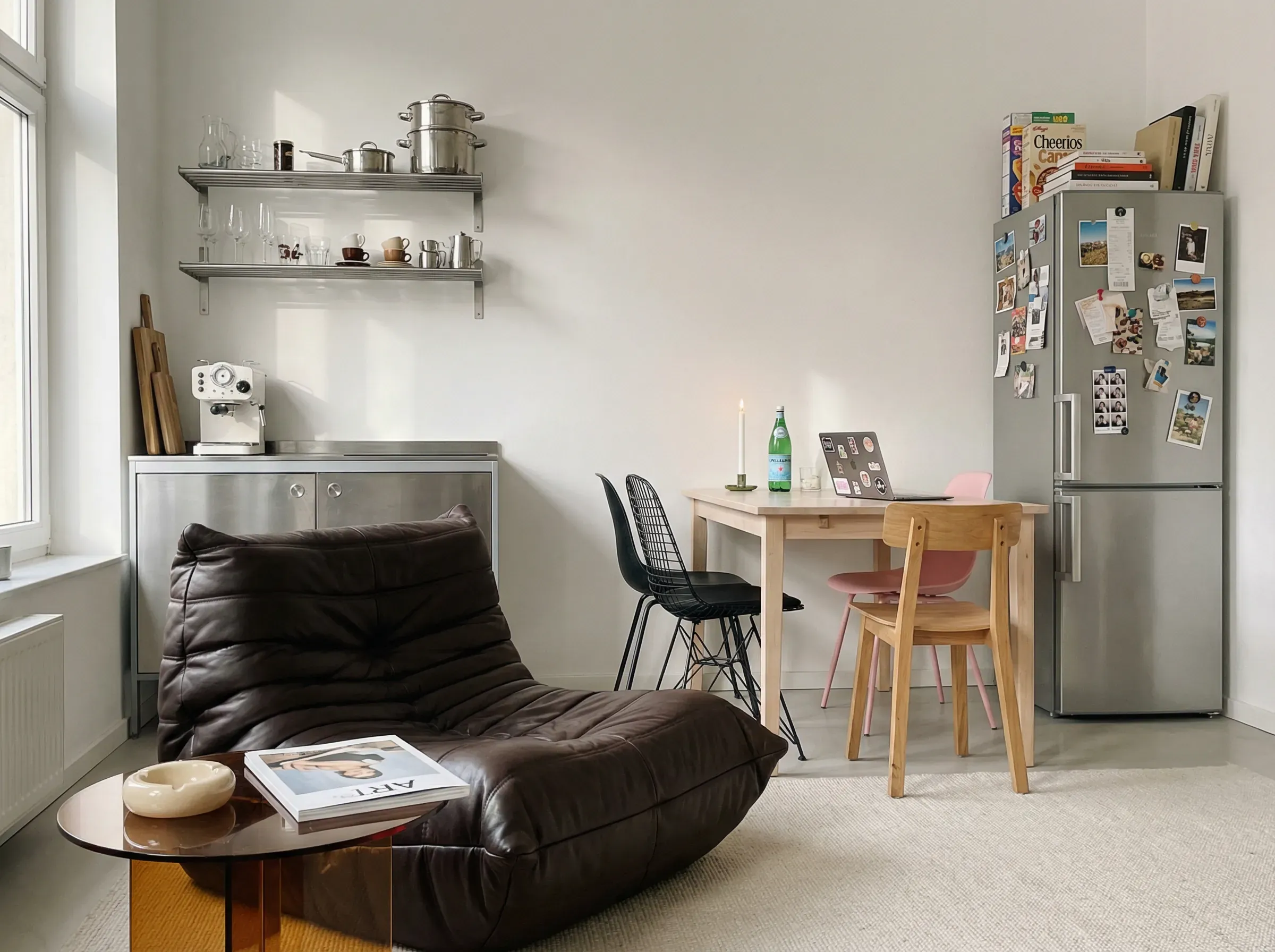 Interior photography of a sunlit eclectic kitchen-living space. 35mm lens, warm late-morning natural light, lightly desaturated colors, airy but intimate atmosphere.
Left: A long white wall with a mounted chrome shelf holding mismatched glasses, a small espresso cup collection, and two stacked stainless steel pots. Beneath it, a narrow counter in brushed steel with a compact white espresso machine and a wooden cutting board leaning vertically.
Foreground Center: A rounded, deeply tufted lounge chair in dark chocolate leather with a slouchy, folded silhouette. Beside it, a low circular side table in translucent amber acrylic with a lighter, ashtray, and folded magazine on top.
Midground: A simple rectangular dining table in pale wood with one white candle, a green bottle of sparkling water, and an open laptop covered in small stickers. Three chairs around it, all mismatched: one black wire chair, one pink molded plastic chair, one unfinished wood chair.
Right: A freestanding silver fridge with a collage of postcards, receipt scraps, magnets, and one strip of photobooth portraits. Above it are stacked cereal boxes and art books. Pale rug, soft shadows, editorial lived-in apartment mood.